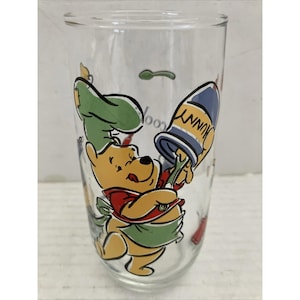 May include: Clear glass tumbler featuring a cartoon illustration of Winnie the Pooh in a chef's outfit. The design includes the word "HUNNY" on a blue and yellow container. The glass has a tall, cylindrical shape and is decorated with colorful graphics.