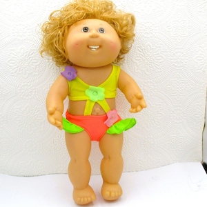 May include: A doll with curly blonde hair and a round face wearing a yellow top with a purple flower, and orange bottoms with green ruffles and a pink flower. The doll has a light skin tone, dark eyes, and a small open mouth showing teeth.