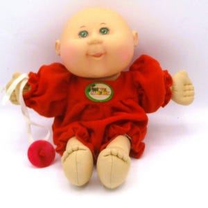 May include: A vintage Cabbage Patch Kids doll with a bald head, green eyes, and rosy cheeks. The doll wears a red dress with a green and yellow patch. It has beige hands and feet, and a red ball attached to a white string.