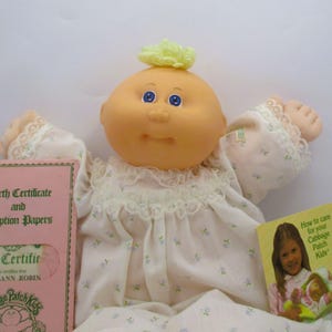 May include: A Cabbage Patch Kids doll with a round peach-colored face, blue eyes, and a small tuft of yellow yarn hair. The doll wears a white long-sleeved gown with a small blue floral print and lace trim. Includes a pink birth certificate folder.