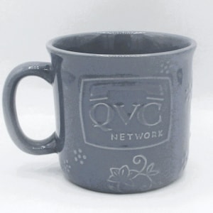 May include: A gray ceramic mug with a handle. The mug features the text "QVC NETWORK" in a rectangular frame. Decorative floral and dot patterns are also visible on the mug's surface. The mug is designed for beverages.