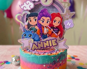 May include: A round cake with horizontal stripes of teal, orange, and pink frosting, decorated with purple glitter and star sprinkles. A large cake topper features the name ANNIE, the number 5, and three cartoon characters with a blue cat.