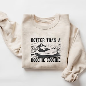 Puede incluir: Sudadera color crema con cuello redondo. La parte delantera presenta un gráfico en blanco y negro de una persona en un flotador en el agua, con el texto "HOTTER THAN A HOOCHIE COOCHIE" en una fuente vintage en negrita.