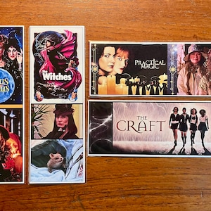 May include: Five different movie posters featuring witches and magic. The posters are all in a vertical format and are arranged in a row. The posters are all on a wooden surface. The posters are for the movies Hocus Pocus, The Witches, Practical Magic, The Craft, and The Craft.