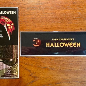 May include: Two stickers featuring the title card from the 1978 horror film "Halloween" by John Carpenter. The first sticker features a black background with the title "Halloween" in white text and a jack-o-lantern with a menacing grin. The second sticker features a black background with the title "Halloween" in white text and the tagline "The Night He Came Home!" with a bloody knife and hand.