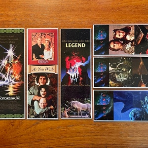 May include: A collection of rectangular bookmarks featuring movie posters. Titles include "Willow", "Excalibur", "The Princess Bride", and "Legend". The bookmarks display various scenes and character portraits from the films, with vibrant colors and detailed imagery.