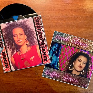 May include: Two 7-inch vinyl records with a black and white photo of a woman with curly hair. The record covers are pink, gold, and black with the text "Buffalo Stance" and "Renee Cherry".