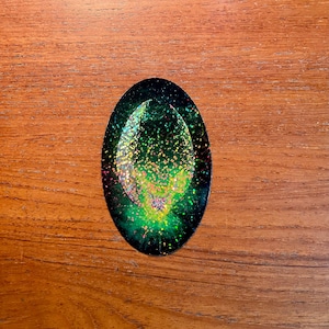 May include: An oval-shaped sticker with a dark green border and a vibrant, glittery interior. The center features a lighter green and yellow design with scattered glitter, creating a sparkling effect. The sticker is set against a wooden background.