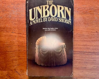 May include: A paperback book cover with a dark background and the title "The Unborn" in large, yellow letters. The subtitle reads "A Novel by David Shobin". Below the title is a black and white illustration of a baby crib with a glowing light inside.