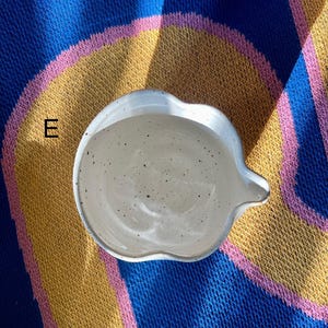 May include: A speckled, off-white ceramic dish with a unique, scalloped edge and a small spout. The dish is set against a colorful background of blue, yellow, and pink fabric.