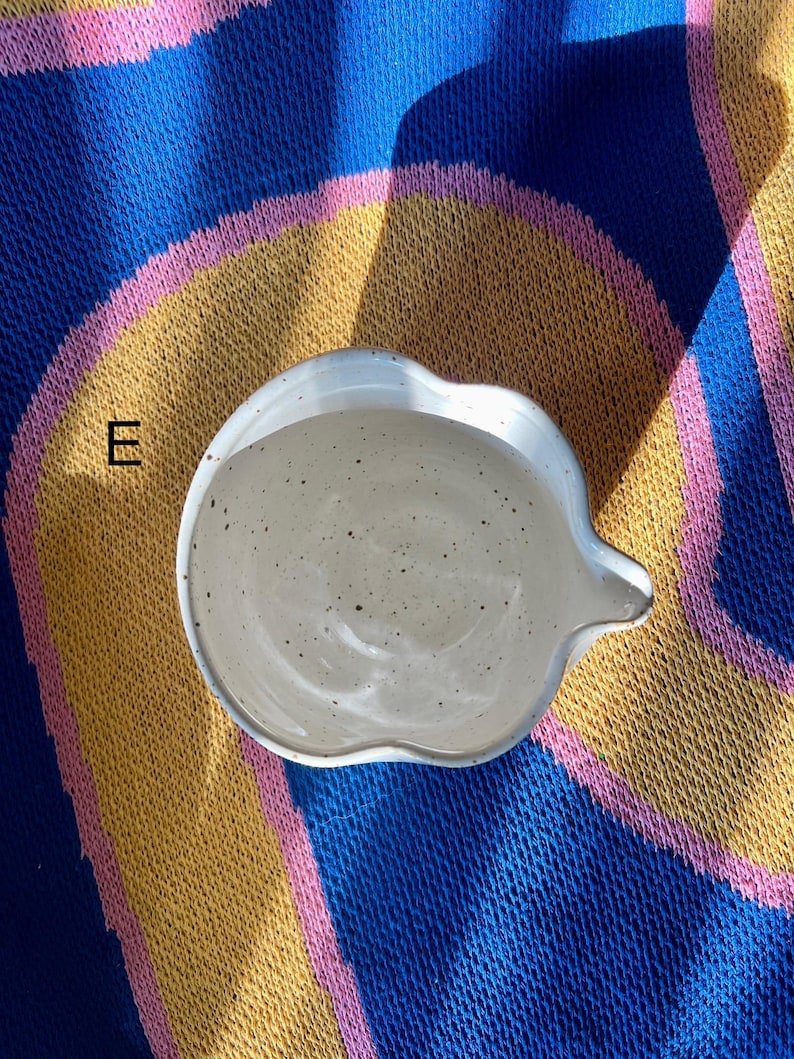 May include: A speckled, off-white ceramic dish with a unique, scalloped edge and a small spout. The dish is set against a colorful background of blue, yellow, and pink fabric.