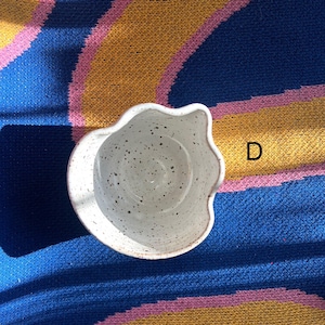 May include: A speckled white ceramic bowl with a wavy rim. The bowl is set against a blue, yellow, and pink patterned background. The letter "D" is visible in the background.