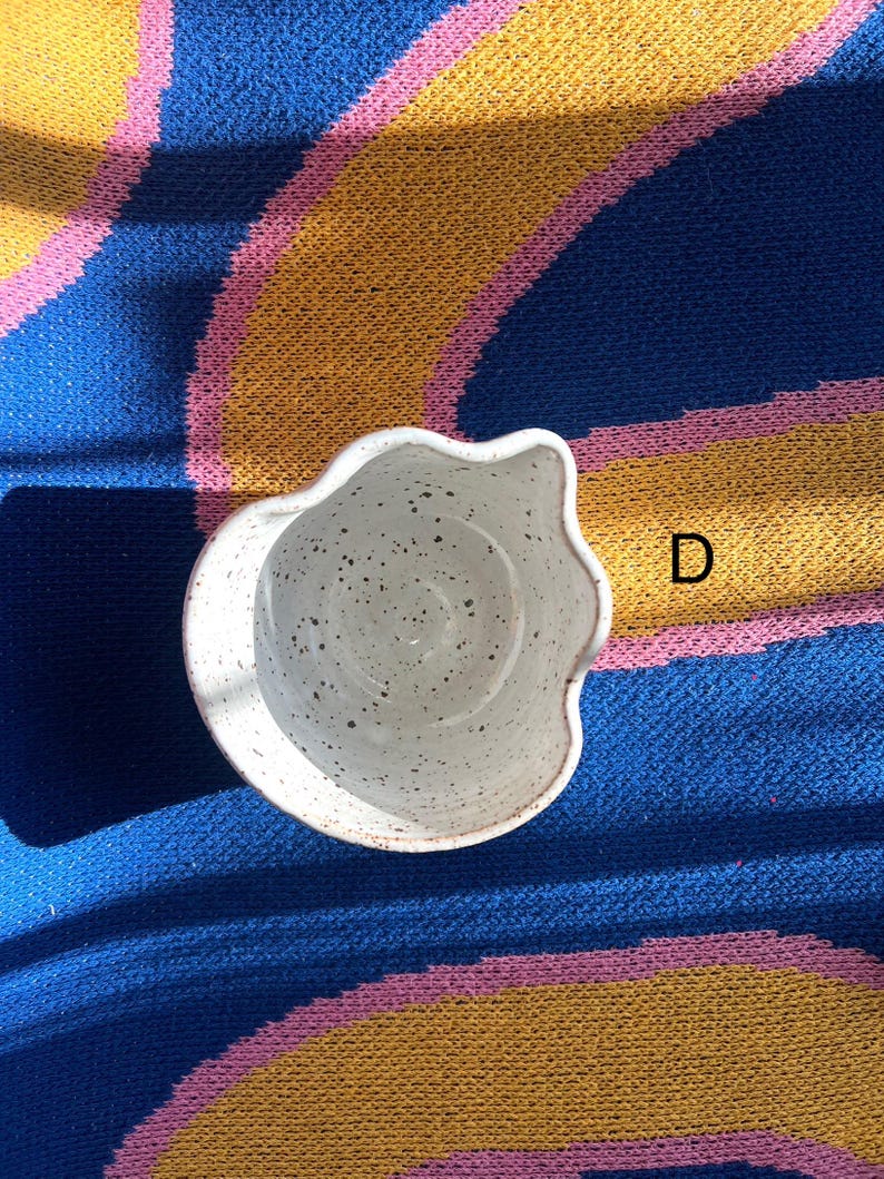 May include: A speckled white ceramic bowl with a wavy rim. The bowl is set against a blue, yellow, and pink patterned background. The letter "D" is visible in the background.