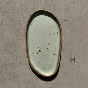May include: An oval-shaped ceramic plate with an irregular, organic edge. The surface features a pale mint green glaze with dark brown speckles and a single reddish-brown fleck. The rim is exposed, showing a textured, raw brown clay finish. The letter H is visible.