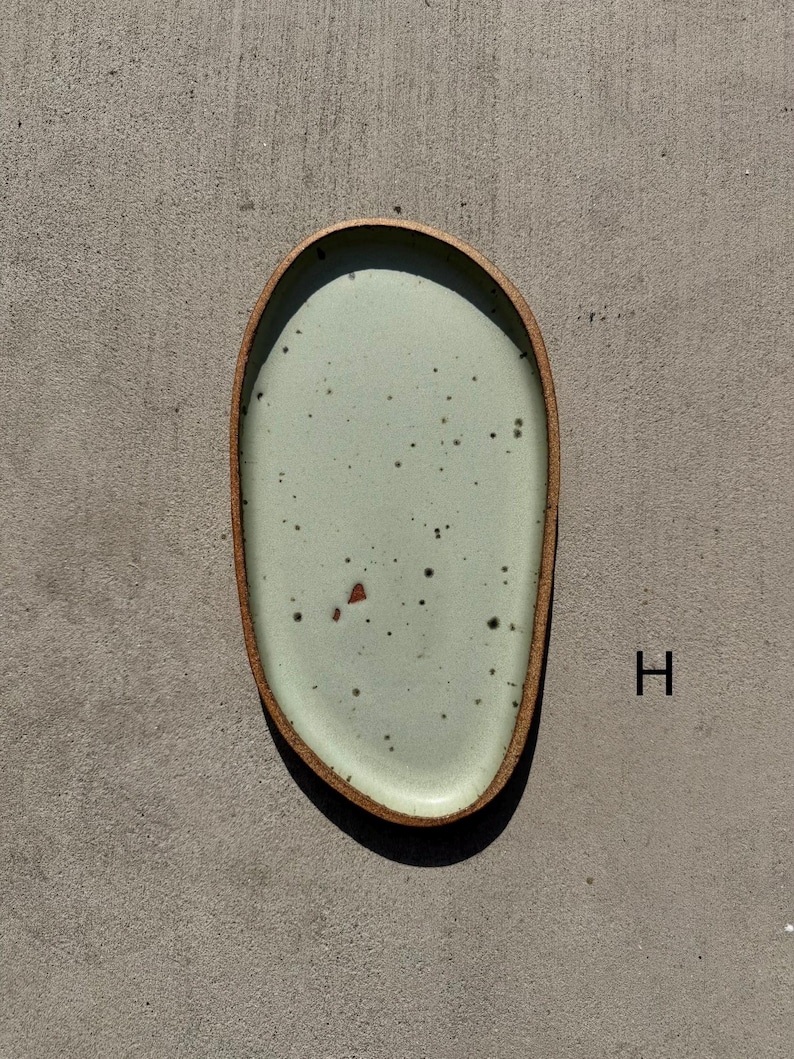May include: An oval-shaped ceramic plate with an irregular, organic edge. The surface features a pale mint green glaze with dark brown speckles and a single reddish-brown fleck. The rim is exposed, showing a textured, raw brown clay finish. The letter H is visible.