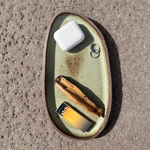May include: An oval-shaped ceramic tray with a light green glazed center and a dark brown rim. The tray holds a white wireless earbud charging case, two silver rings, a piece of light brown wood, and a small matchbox with a yellow gradient.