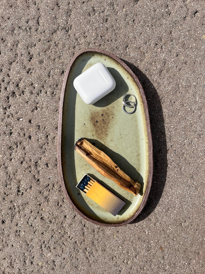 May include: An oval-shaped ceramic tray with a light green glazed center and a dark brown rim. The tray holds a white wireless earbud charging case, two silver rings, a piece of light brown wood, and a small matchbox with a yellow gradient.