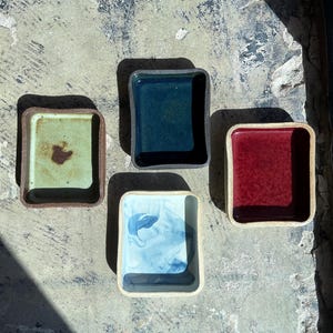 May include: Four small, square ceramic dishes in shades of green, blue, and red. The dishes have a slightly raised rim and a glossy finish. The background is a textured, light-colored surface.