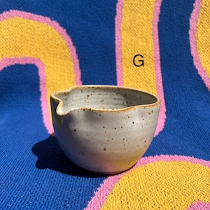 May include: A small, speckled ceramic bowl with a spout, perfect for serving sauces or dips. The bowl is a light gray color with a slightly textured surface and a brown rim. The background is a blue, yellow, and pink patterned textile.