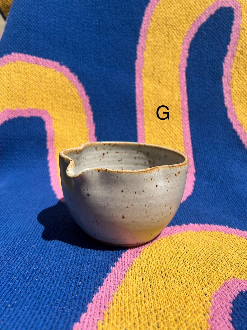 May include: A small, speckled ceramic bowl with a spout, perfect for serving sauces or dips. The bowl is a light gray color with a slightly textured surface and a brown rim. The background is a blue, yellow, and pink patterned textile.