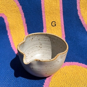 May include: A small, off-white ceramic bowl with a spout and a speckled texture. The bowl has a wavy rim and a light brown edge. The background is a blue, yellow, and pink patterned textile.