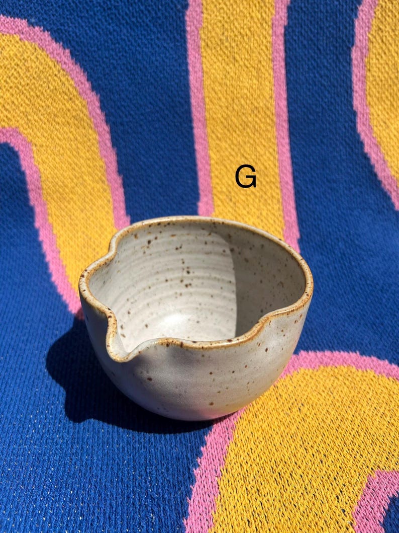 May include: A small, off-white ceramic bowl with a spout and a speckled texture. The bowl has a wavy rim and a light brown edge. The background is a blue, yellow, and pink patterned textile.