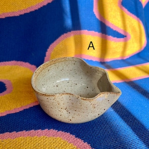 May include: A small, speckled ceramic bowl with a unique, asymmetrical shape. The bowl is a light beige color with brown speckles and a slightly wavy rim. The bowl has a spout on one side. The background is a blue, yellow, and pink patterned fabric.