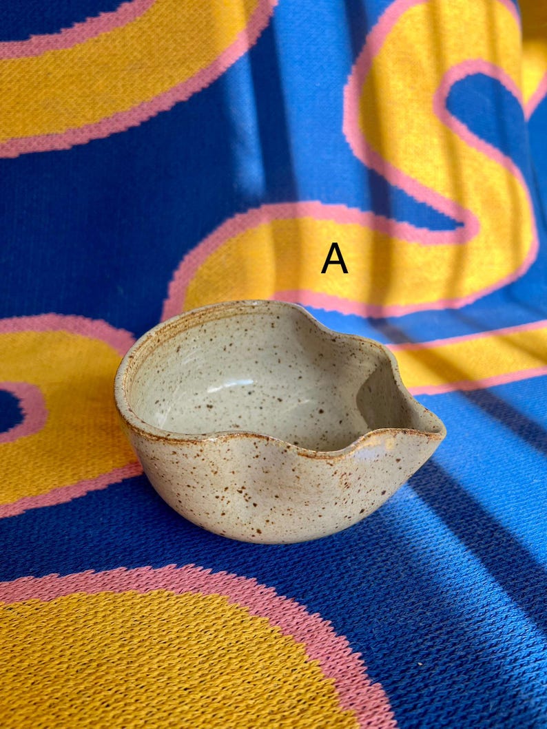 May include: A small, speckled ceramic bowl with a unique, asymmetrical shape. The bowl is a light beige color with brown speckles and a slightly wavy rim. The bowl has a spout on one side. The background is a blue, yellow, and pink patterned fabric.