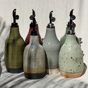 May include: A collection of ceramic oil bottles in various colors, including green, gray, and speckled white. Each bottle has a black spout and a dark base. The bottles are arranged on a neutral-colored surface.