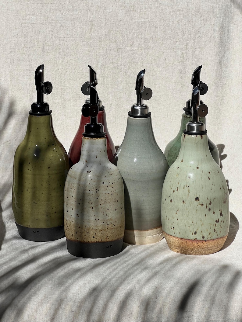 May include: A collection of six ceramic oil dispensers with varied glaze finishes including olive green, speckled cream, light blue-grey, and sage green. Each bottle features a black or metallic pour spout with a hinged lid and a rounded base.