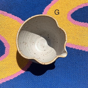 May include: A speckled, off-white ceramic bowl with a spout. The bowl has a unique, organic shape and a brown rim. The bowl is sitting on a blue, yellow, and pink striped surface.
