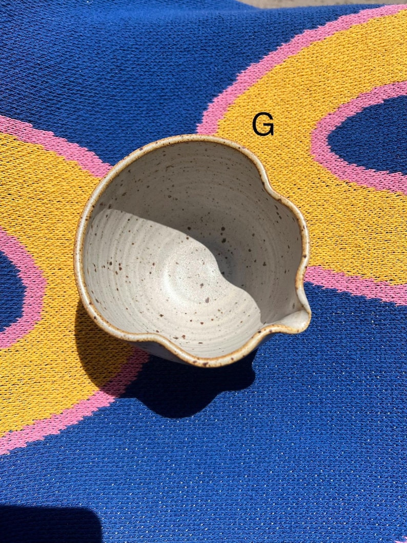 May include: A speckled, off-white ceramic bowl with a spout. The bowl has a unique, organic shape and a brown rim. The bowl is sitting on a blue, yellow, and pink striped surface.