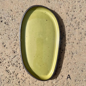 May include: An oval-shaped ceramic tray with a smooth, glossy light green interior surface featuring subtle dark speckles. The tray has a dark, textured, matte-finished outer rim and edge. The letter A is printed in black near the bottom right.