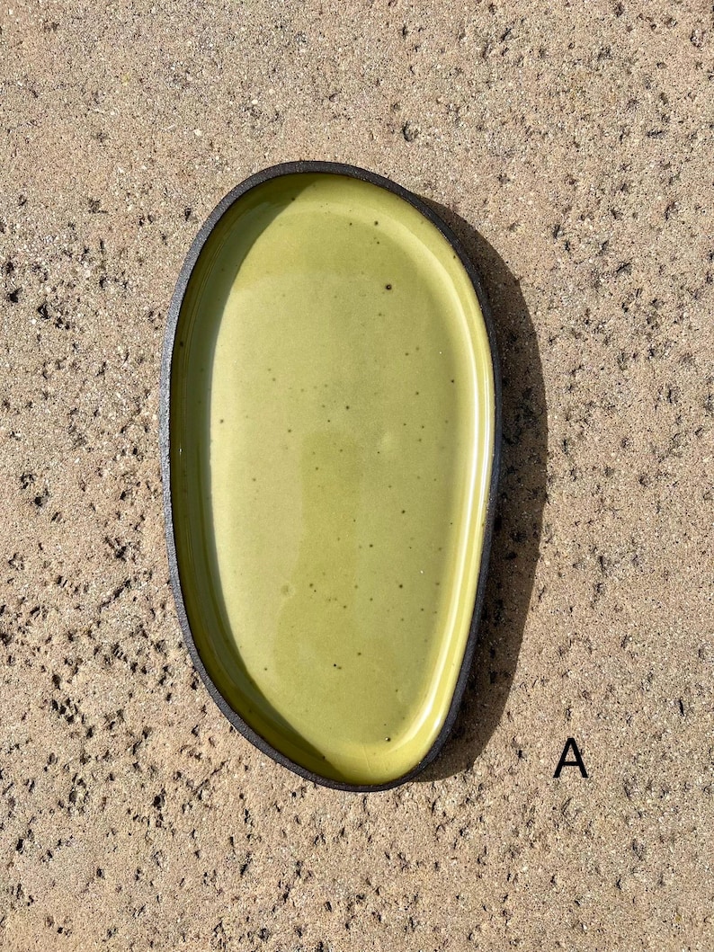 May include: An oval-shaped ceramic tray with a smooth, glossy light green interior surface featuring subtle dark speckles. The tray has a dark, textured, matte-finished outer rim and edge. The letter A is printed in black near the bottom right.