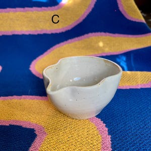 May include: A small, off-white ceramic bowl with a spout. The bowl has a speckled texture and a unique, organic shape. The background is a blue, yellow, and pink patterned textile.