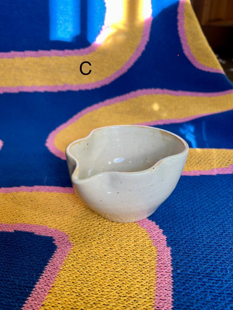 May include: A small, off-white ceramic bowl with a spout. The bowl has a speckled texture and a unique, organic shape. The background is a blue, yellow, and pink patterned textile.