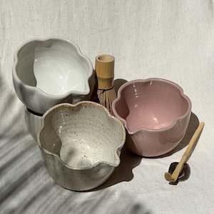 May include: A selection of ceramic matcha bowls in white, speckled, and pink. The bowls feature a distinctive scalloped edge design. A bamboo whisk, a bamboo scoop, and a small black ceramic rest are also included.