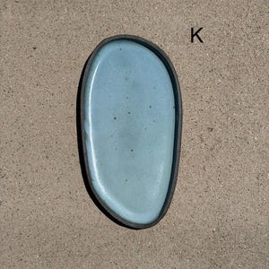May include: An oval-shaped ceramic tray with a smooth, light blue glazed interior surface featuring subtle dark speckles. The tray has a dark, matte-finished outer rim and edge. The letter K is printed in black near the upper right side of the tray.