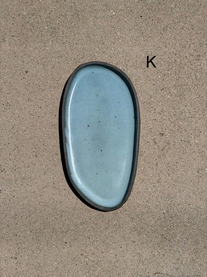 May include: An oval-shaped ceramic tray with a smooth, light blue glazed interior surface featuring subtle dark speckles. The tray has a dark, matte-finished outer rim and edge. The letter K is printed in black near the upper right side of the tray.