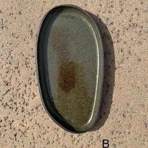 May include: An oval-shaped ceramic tray with a dark, textured outer rim and a glossy, mottled olive-green interior glaze. The center features a darker brown, speckled patch. The letter B is printed in black near the bottom right corner.