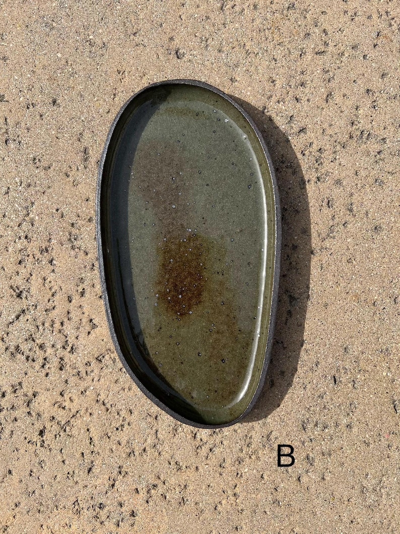 May include: An oval-shaped ceramic tray with a dark, textured outer rim and a glossy, mottled olive-green interior glaze. The center features a darker brown, speckled patch. The letter B is printed in black near the bottom right corner.