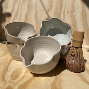 May include: A collection of handmade ceramic bowls in various sizes and shapes, with a light beige and speckled blue-gray glaze. A bamboo whisk sits beside the bowls. The bowls have organic, wavy rims and are arranged on a light wood surface.