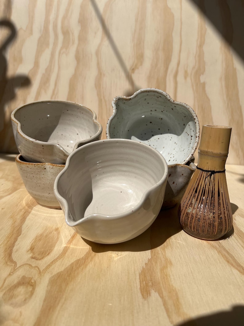 May include: A collection of handmade ceramic bowls in various sizes and shapes, with a light beige and speckled blue-gray glaze. A bamboo whisk sits beside the bowls. The bowls have organic, wavy rims and are arranged on a light wood surface.