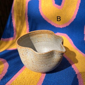 May include: A speckled, light-colored ceramic bowl with a wavy rim and a small spout. The bowl is set against a colorful blue, yellow, and pink patterned background. The letter "B" is visible on the background.