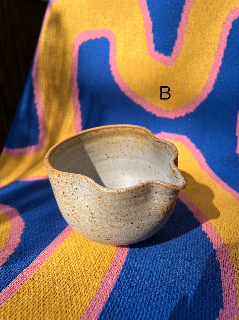 May include: A speckled, light-colored ceramic bowl with a wavy rim and a small spout. The bowl is set against a colorful blue, yellow, and pink patterned background. The letter "B" is visible on the background.