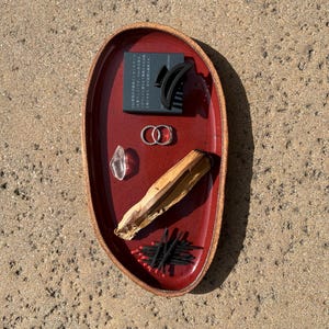 May include: An oval-shaped, deep red ceramic tray with a textured brown rim. The tray holds a black rectangular box with Japanese text, a black hair claw clip, two silver rings, a clear crystal, a piece of light brown wood, and black matches.