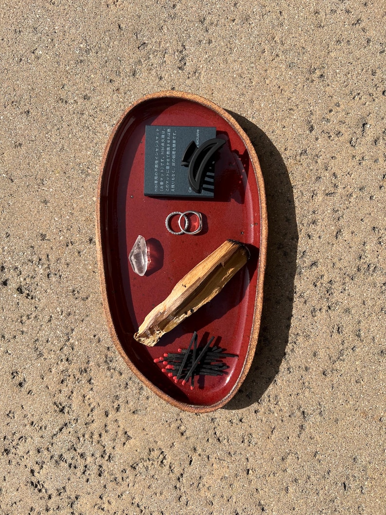 May include: An oval-shaped, deep red ceramic tray with a textured brown rim. The tray holds a black rectangular box with Japanese text, a black hair claw clip, two silver rings, a clear crystal, a piece of light brown wood, and black matches.