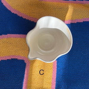 May include: A small, off-white ceramic dish with an irregular, scalloped edge and a spout. The dish is set against a blue, yellow, and pink patterned background. The letter "C" is visible.