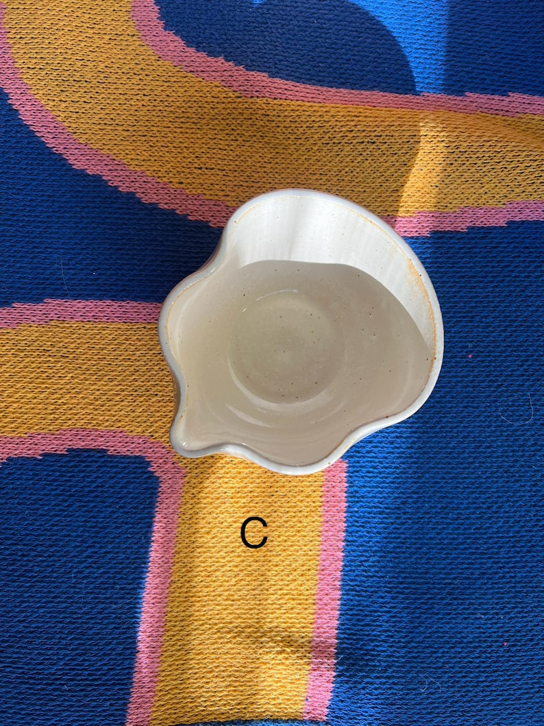 May include: A small, off-white ceramic dish with an irregular, scalloped edge and a spout. The dish is set against a blue, yellow, and pink patterned background. The letter "C" is visible.