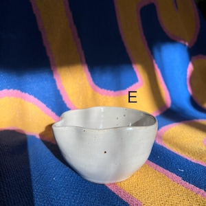 May include: A small, off-white ceramic bowl with a wavy rim. The bowl has a speckled appearance and a small spout. The background is a blue, yellow, and pink patterned fabric.